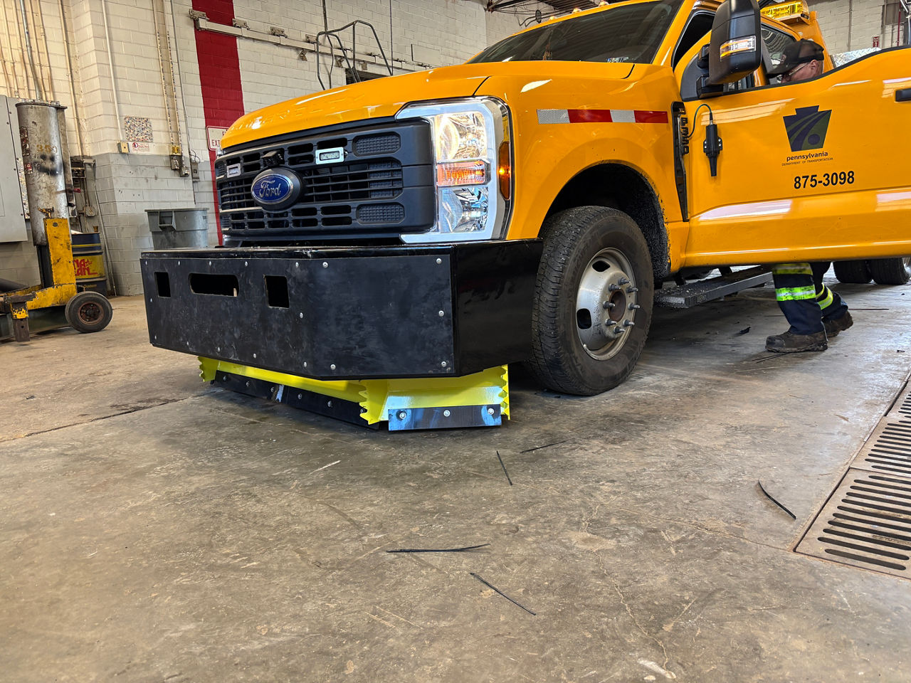 A side view of a yellow truck with a large piece of black metal affixed to the front sitting inside a garage. 