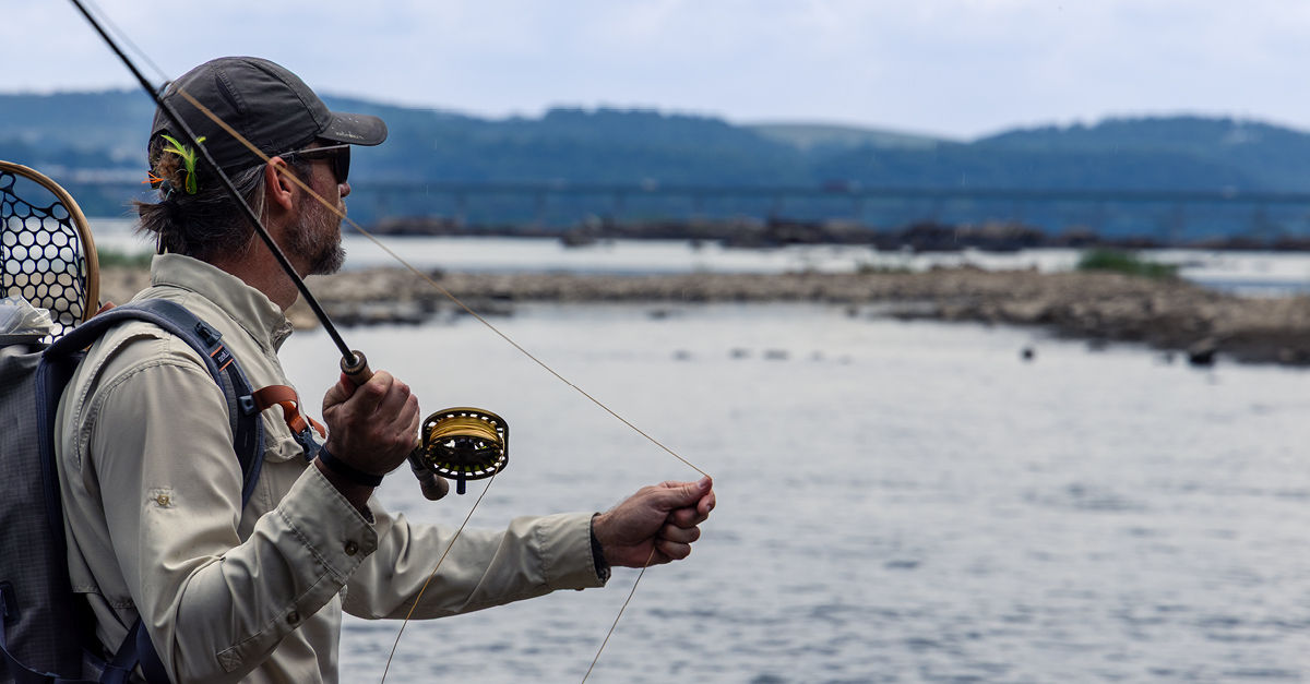 Side profile of a man waist up fly fishing along the Susquehanna River with a scenic mountain and bridge backdrop