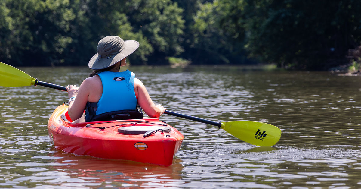 Back of a woman wearing a hat, a life jacket and holding a paddle while kayaking along the Conestoga River
