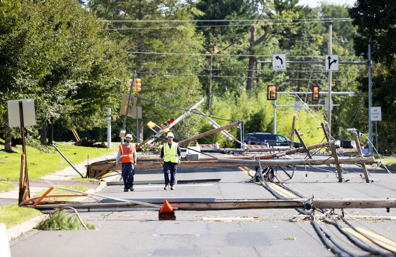Two men in neon safety vests walk among a street full of downed telephone poles. 