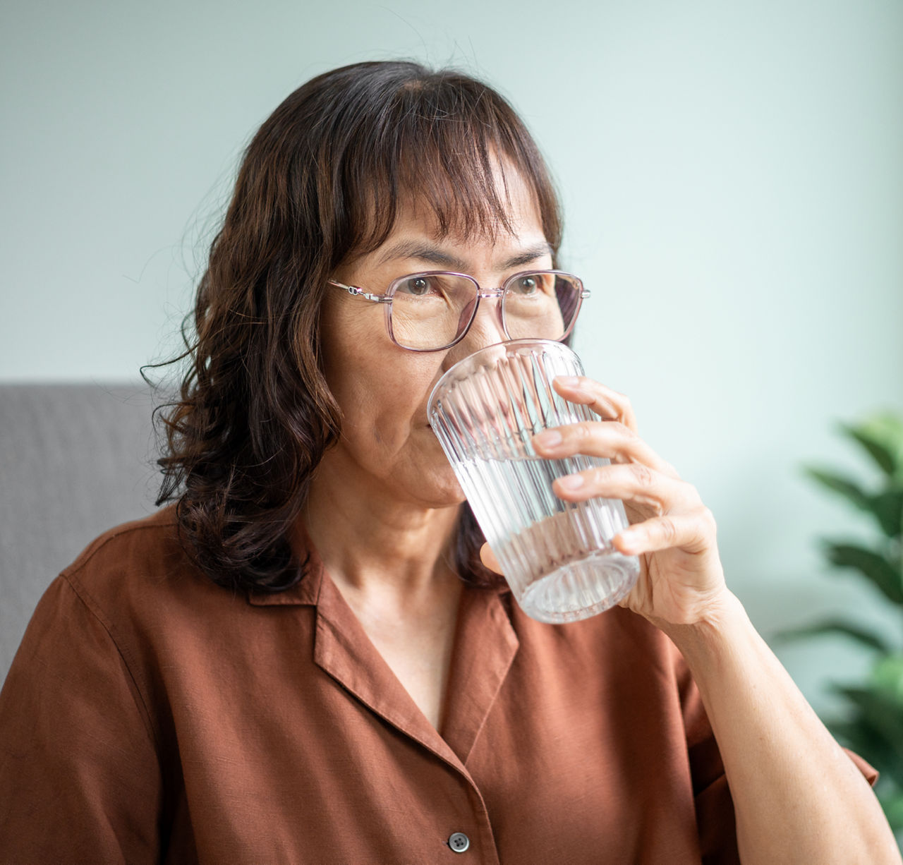 An older woman with shoulder-length hair, bangs, and glasses, sips water on her couch.