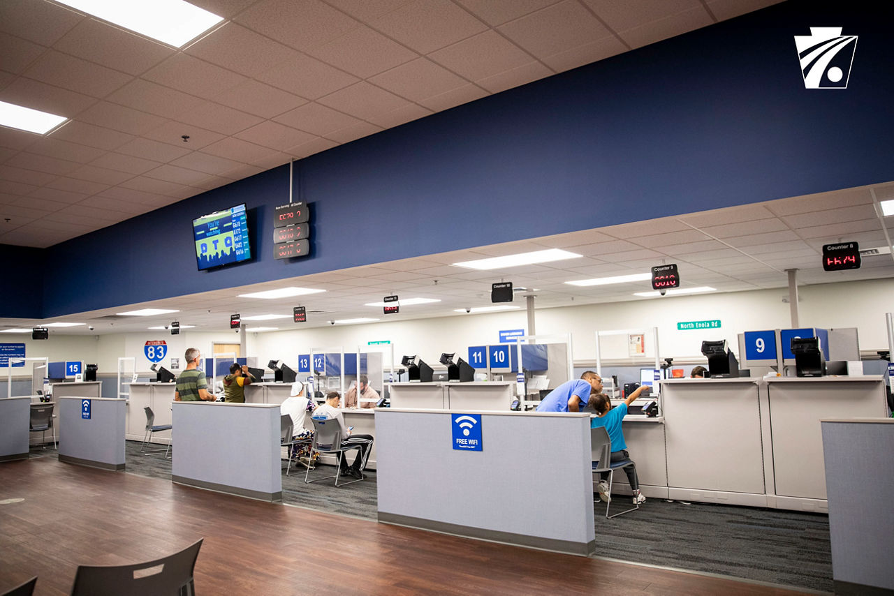 Driver license center with counters for service. Staff are seated behind the counter and customers are seated and standing in front of the counter. There are number signs above the counter.
