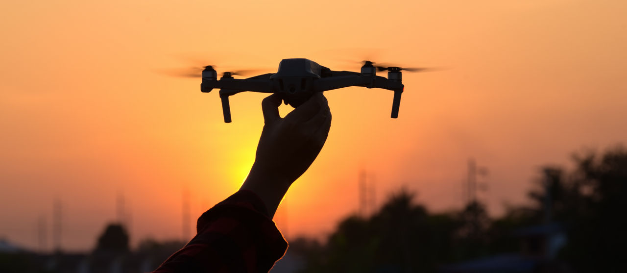 A hand holds up a drone in front of an orange sunset.