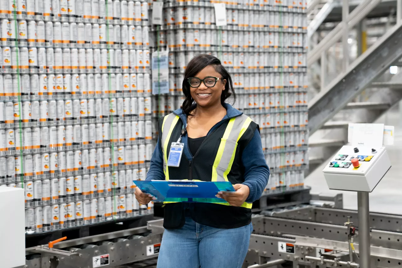 A woman in a safety vest, standing in front of a large palette of canned beverages in a manufacturing facility, smiles.