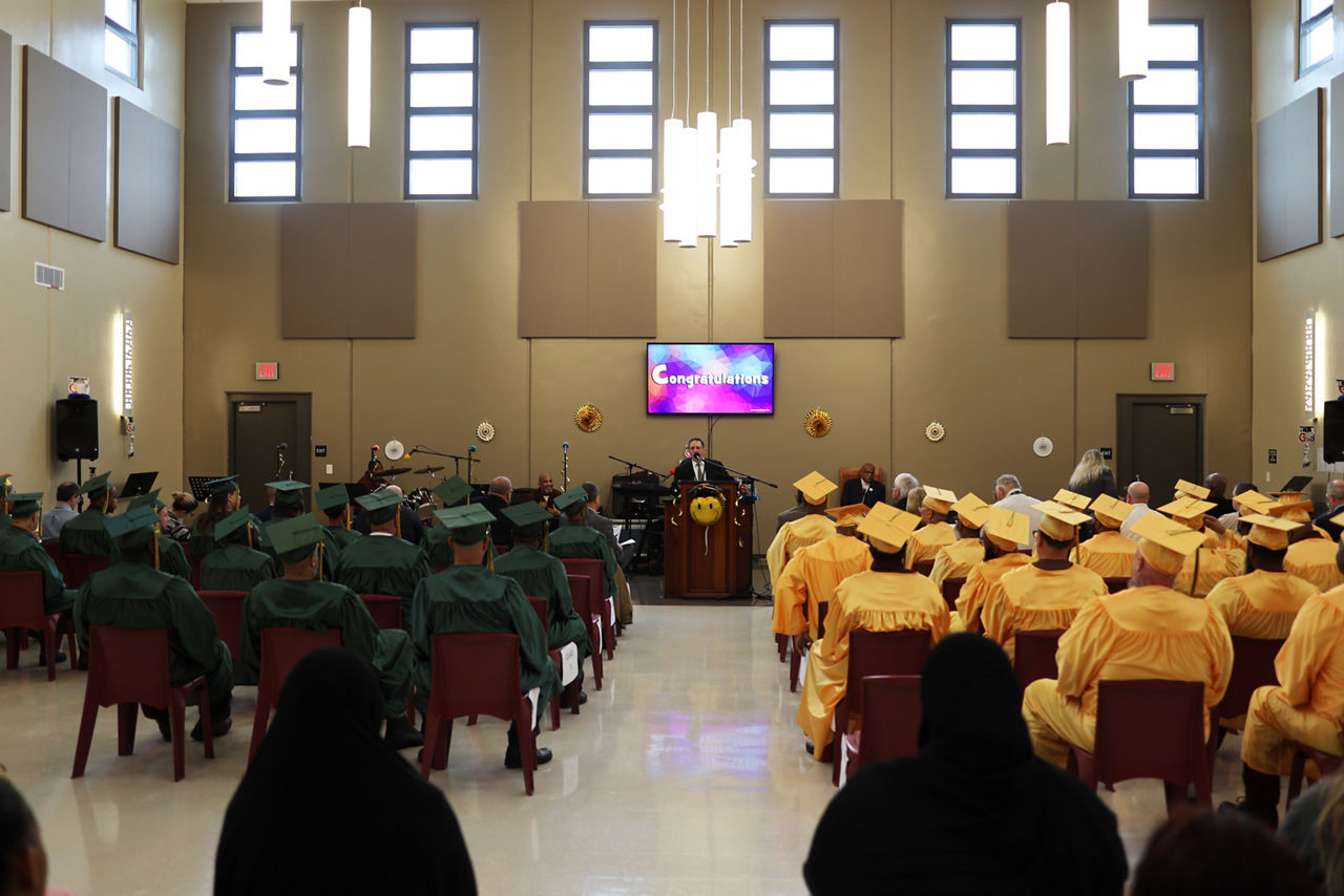 Incarcerated individuals in caps and gowns listen to a speaker during their graduation ceremony