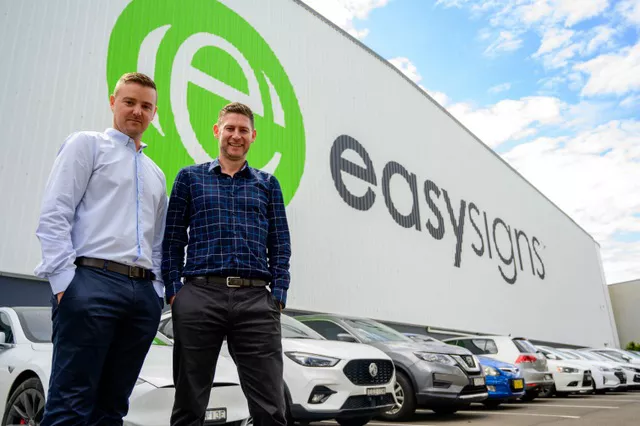 Two men smile for a picture, as they stand in front of a warehouse with the word "Easysigns" printed on the exterior of the building
