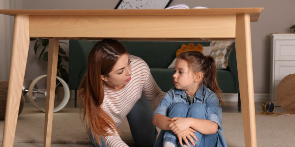 A mother and daughter prepare for an earthquake by seeking protection under a sturdy table.