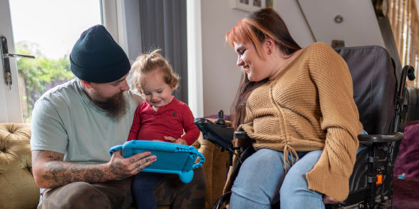 A mom in a wheelchair and a dad sitting on a couch play with a toy with their toddler.