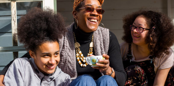 A family smiles. A woman in the center drinks a hot beverage out of a mug and wears sunglasses.