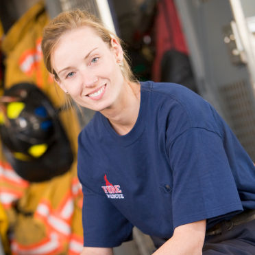 A blond, female firefighter wearing a blue fire department t-shirt smiles at the camera with her hands folded on her knees.