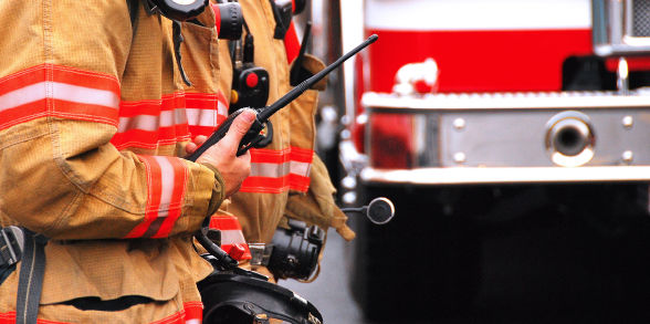 Two firefighters with radios stand near a firetruck.