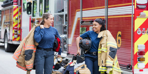 Two female firefighters carry their jackets and gear while smiling at each other.