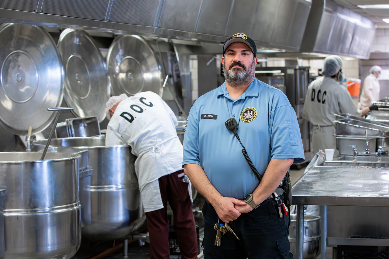 A food service instructor stands in a kitchen while incarcerated individuals work behind him.