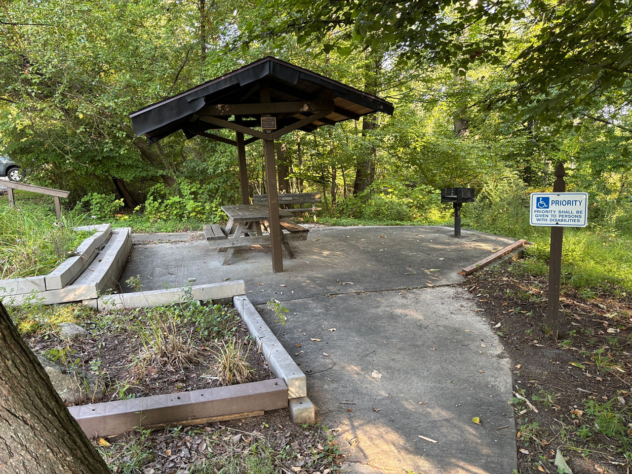 A short but winding sidewalk leads to a small brown pavilion that covers one picnic table and one bench in a wooded area.