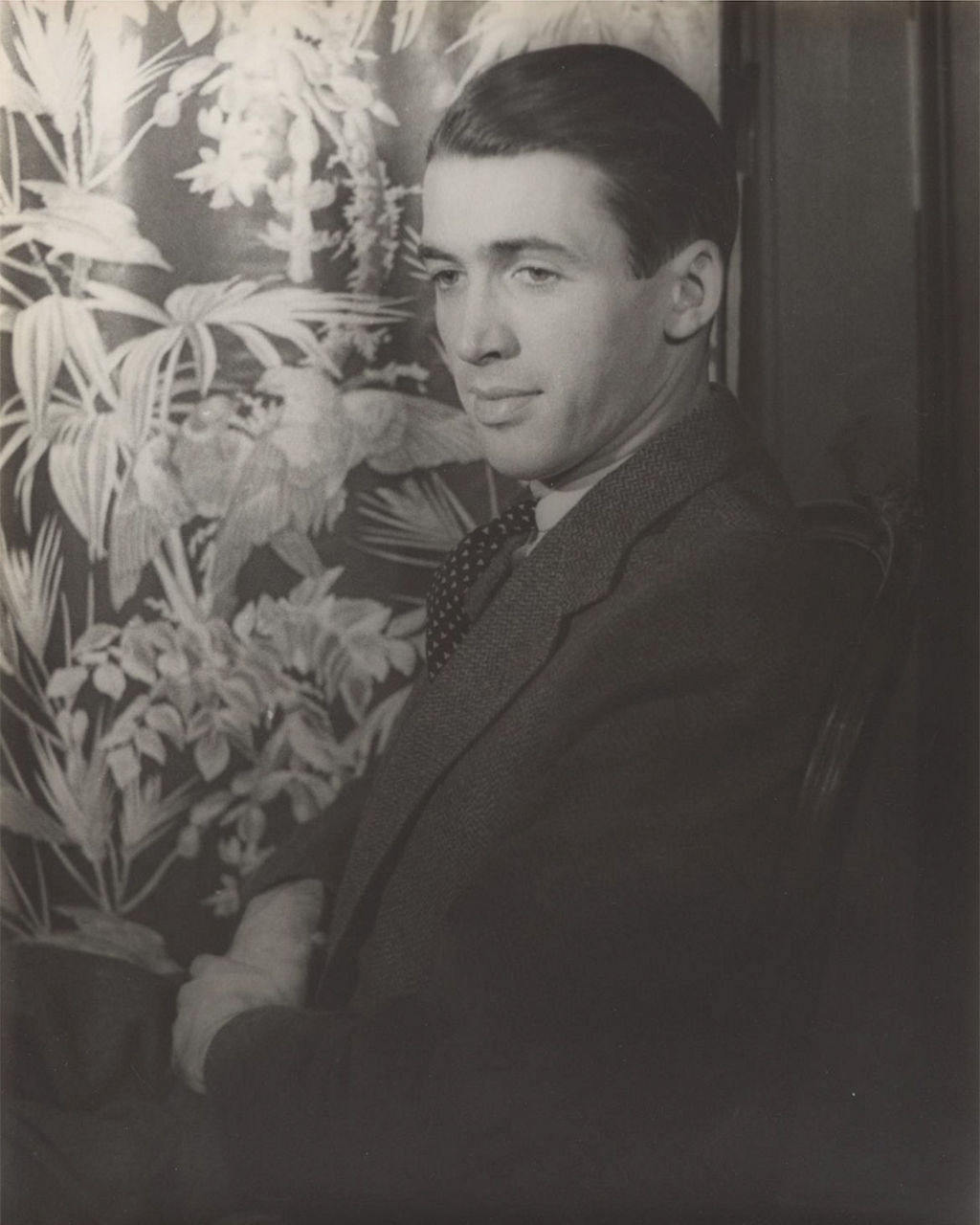 Man in suit seated beside floral wallpaper backdrop