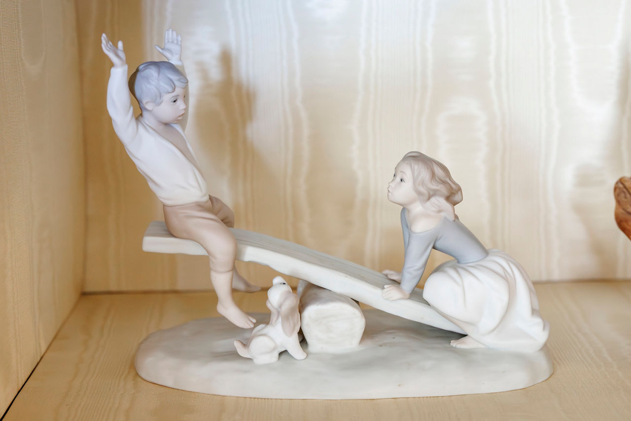 Porcelain figurine of a boy and girl on a seesaw, with a playful dog beneath. 