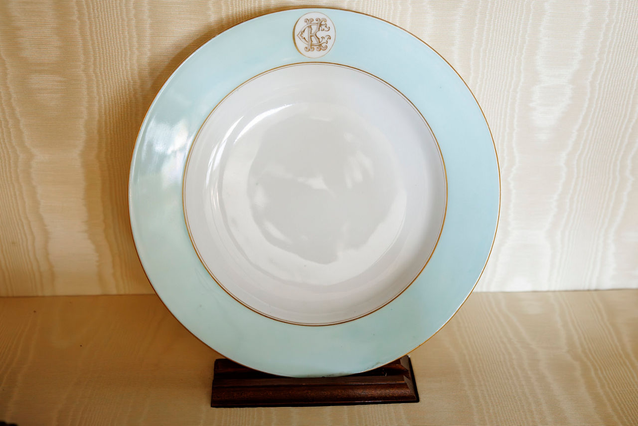 A decorative white plate with a light blue rim and gold trim, featuring an ornate monogram near the top. 