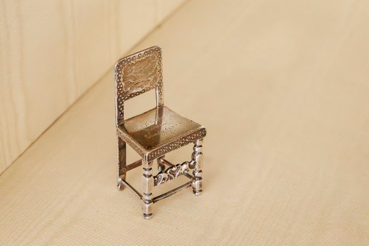 A small, decorative silver chair. It features ornate carvings on the backrest and legs.