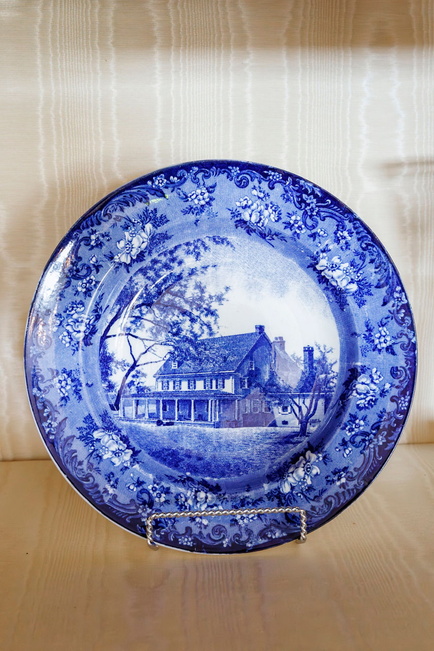 A decorative blue and white porcelain plate displays a detailed rural house scene with surrounding trees. 