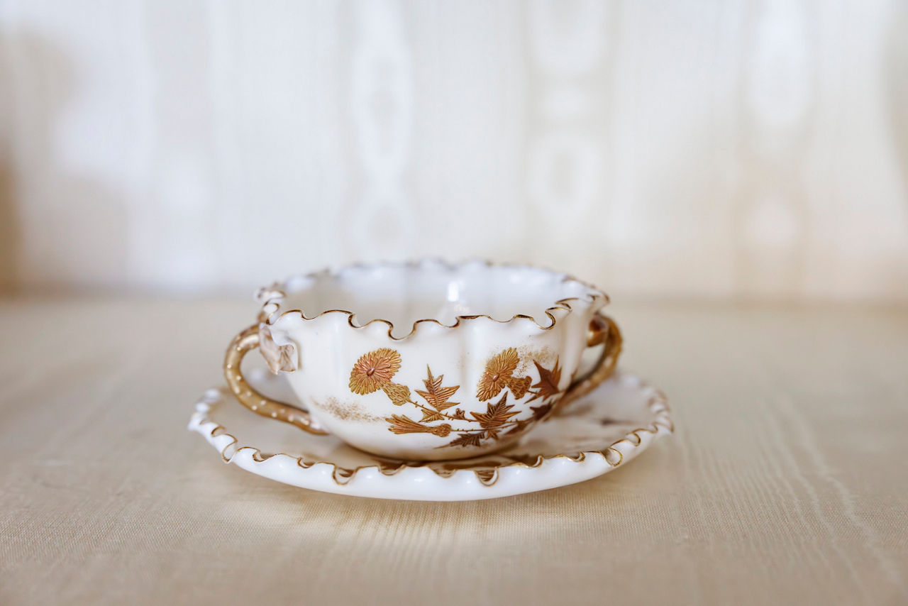 A delicate porcelain teacup with gold floral designs sits on a matching saucer. The scalloped edges add an elegant, antique feel.
