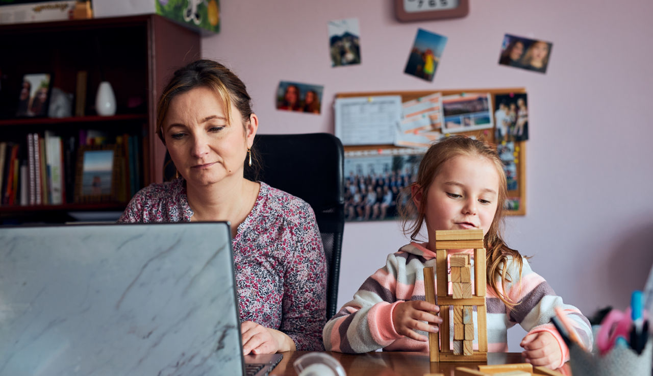 A mom works on her laptop while her daughter stacks wooden blocks.