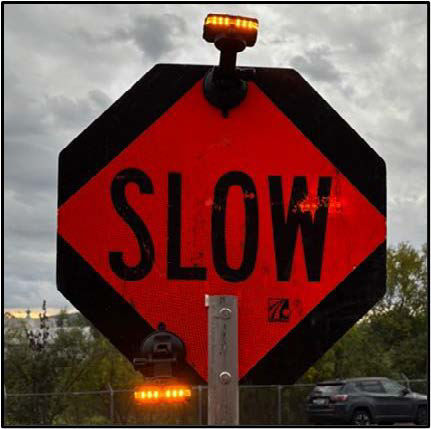 An image of a black and orange sign with the word slow shown in all capital letters on the orange part of the sign. At the top and bottom of the sign, a LED flashing lights are visible.