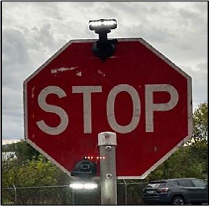 An image of a red sign with white lettering with the word stop shown in all capital letters on the red part of the sign. At the top and bottom of the sign, a LED flashing lights are visible.  