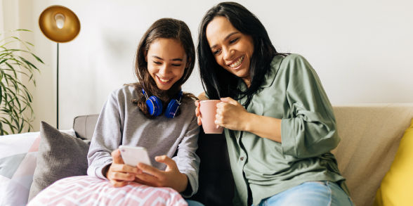 A mother and daughter smile while looking at a cellphone on their couch.