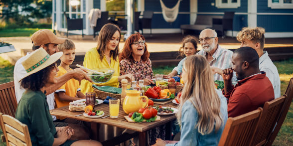 A large family happily eats a meal outside at a picnic table.