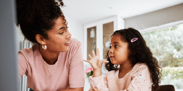 A mom wearing her hair in a curly ponytail talks with her young daughter.