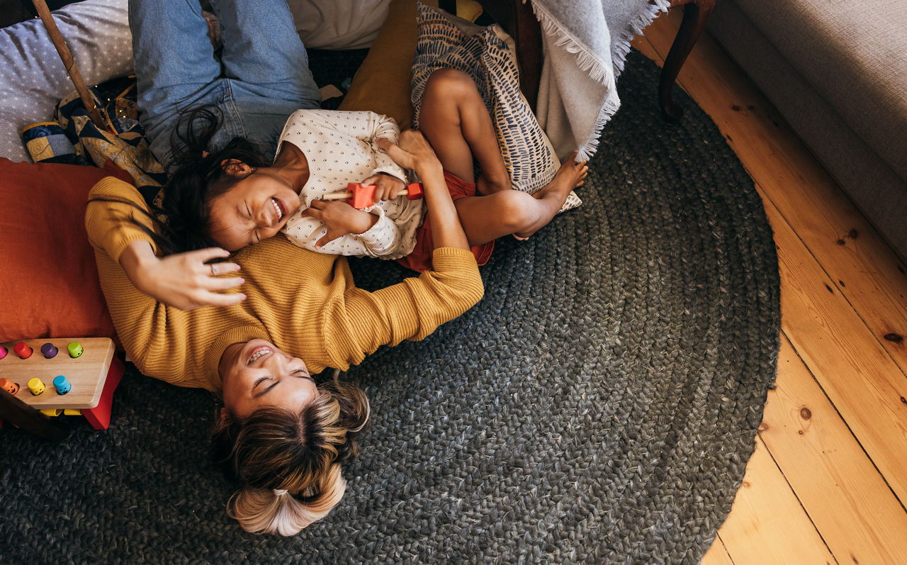 A mom and daughter laugh together on the floor of a cozy room.