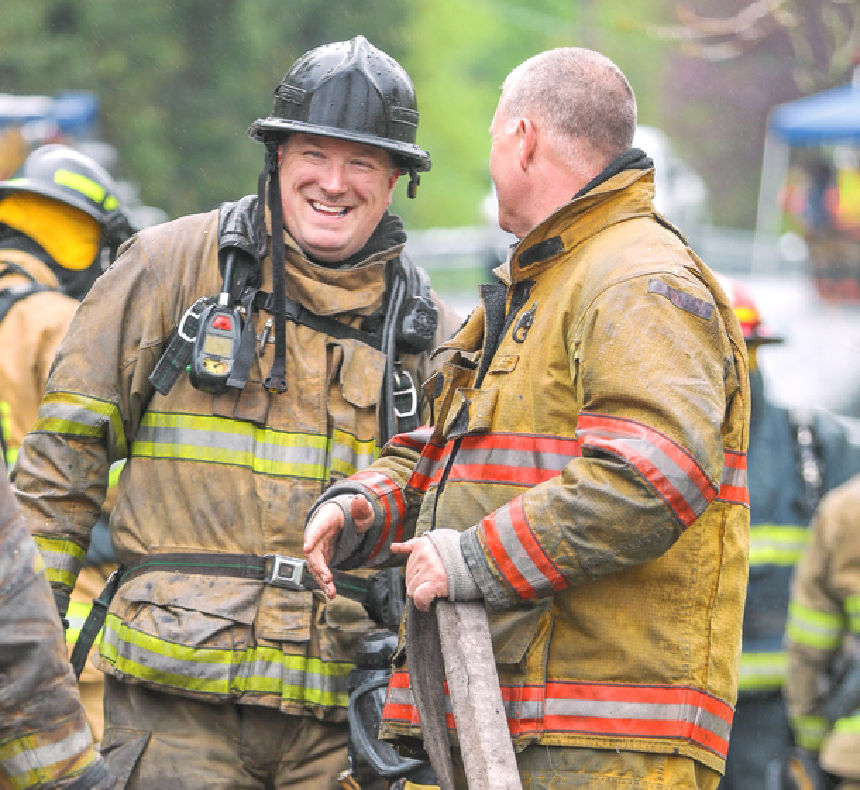 A firefighter smiles outside while chatting with another firefighter.