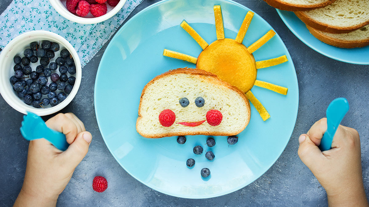 A child sitting in front of a plate of food where the items form a sun and smiley face.