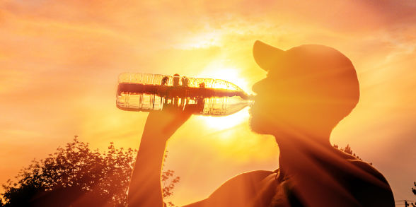 A man with a bright sun behind him wears a hat and chugs from a water bottle.