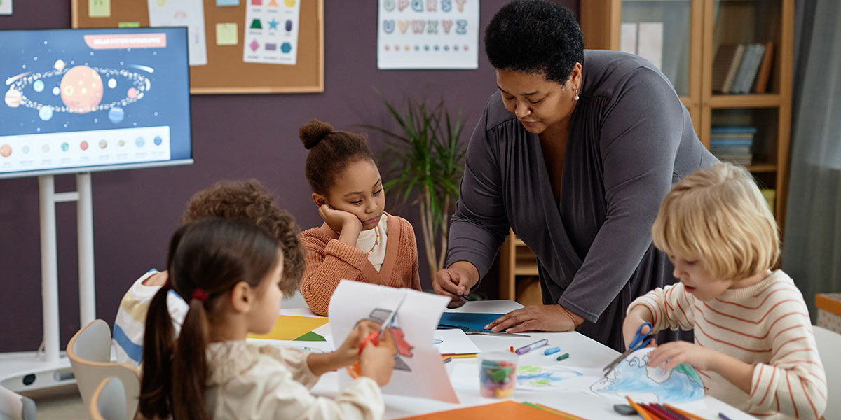 A kind Black Woman helping group of little kids sitting at table in preschool class.