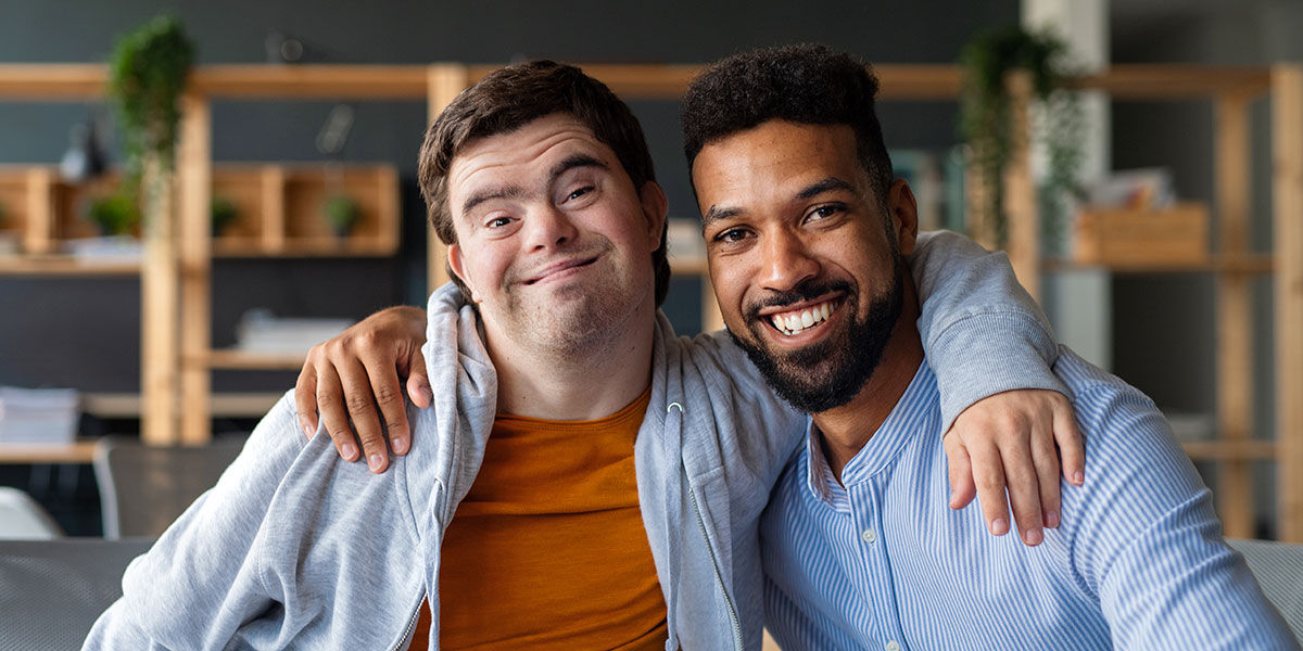 A man with intellectual disability and his support counselor. They both have their arm on each other's shoulder and are smiling at the camera.