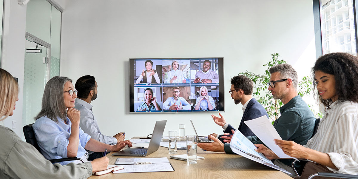 People having a mix of in-person and online meeting with a conference video call on tv screen.
