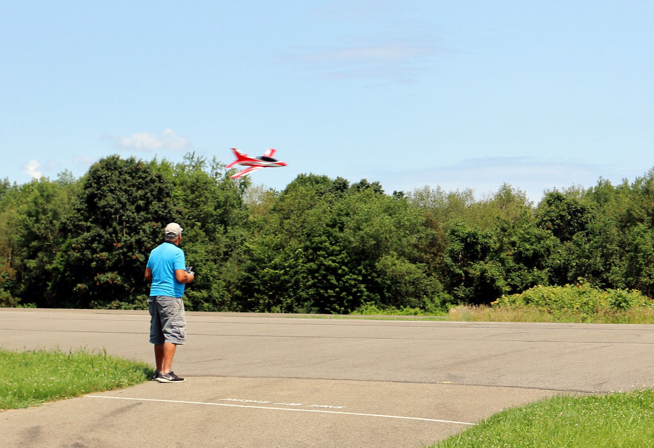 A man radio flying a red model airplane near a green forest