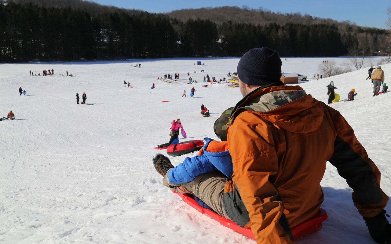 A man sledding down a steep hill with many other sledders enjoying the snowy day