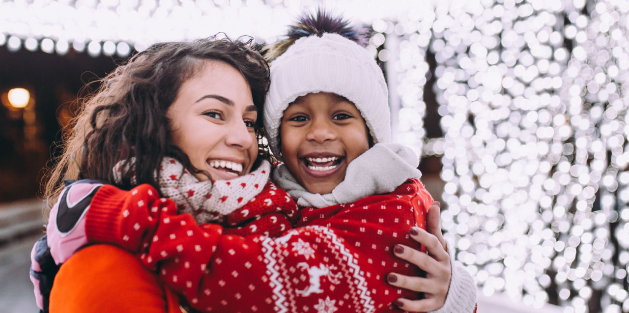 A woman cuddles close to a young girl underneath bright Christmas lights outside.