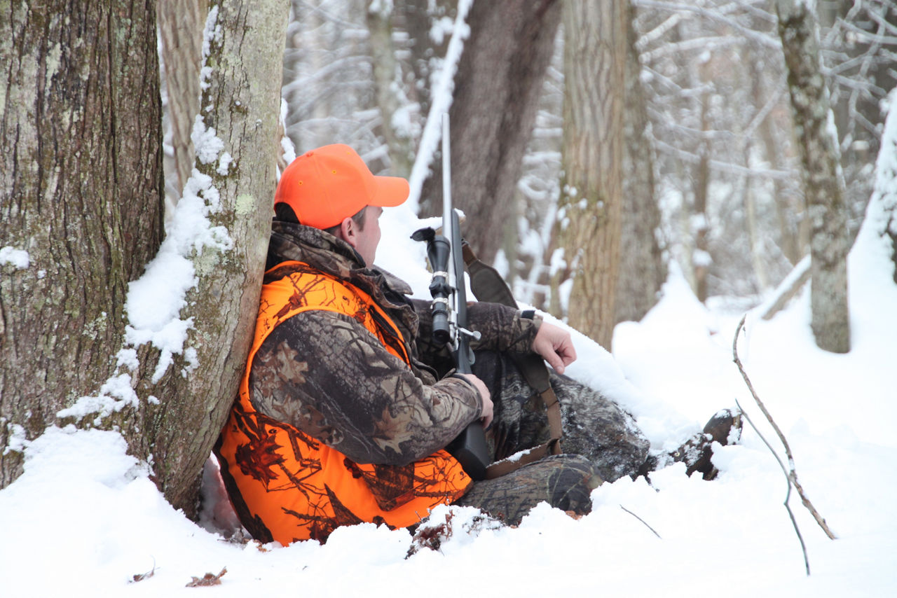 A man wearing orange camouflage clothing is sitting in the snow and leaning against a tree with his gun.