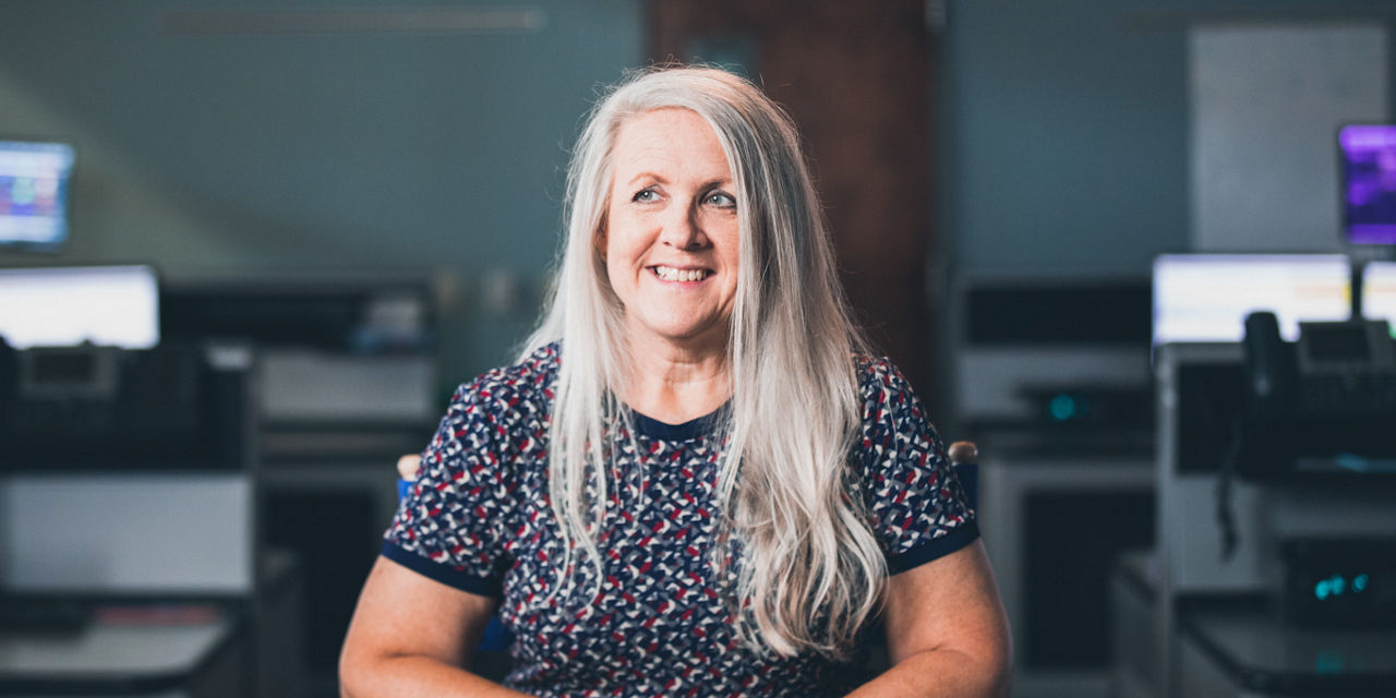 911 dispatcher Lisa sits in a call center and smiles while looking off camera. She has blue eyes and long gray hair, and is wearing a red, white, and blue patterned shirt.