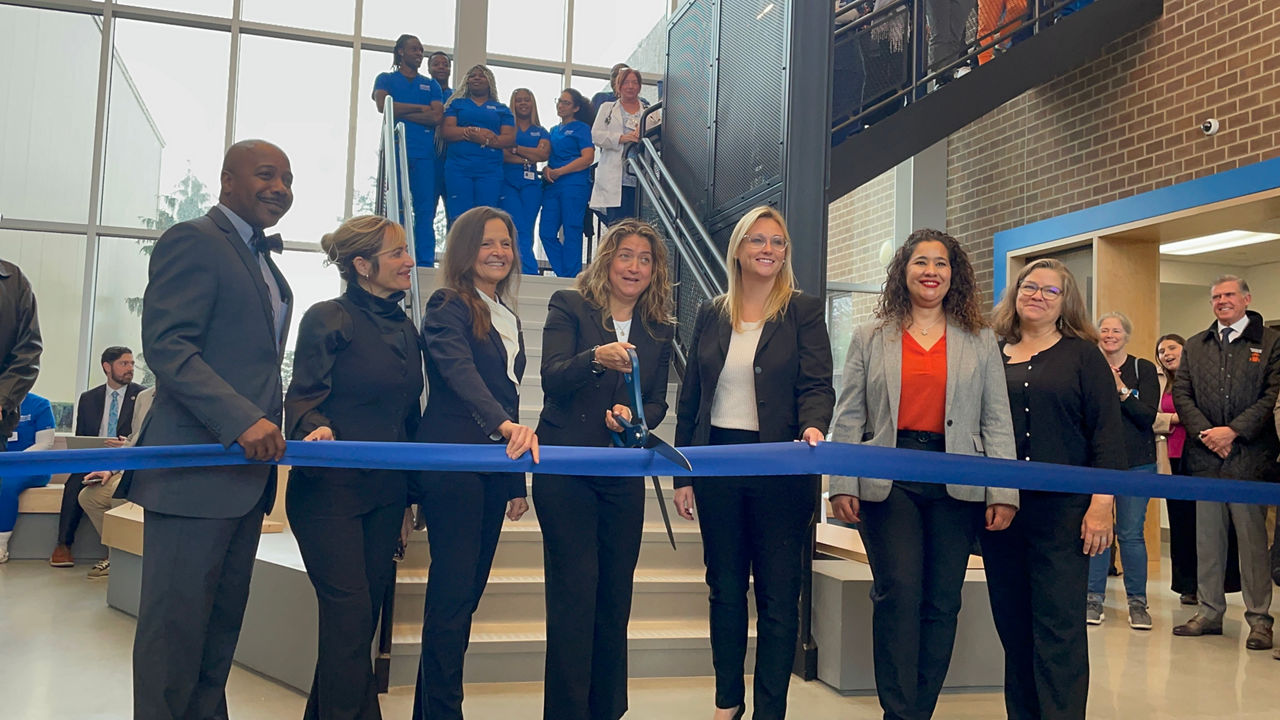 A group of people at a ribbon-cutting ceremony holding a blue ribbon, with one person cutting it with large scissors.