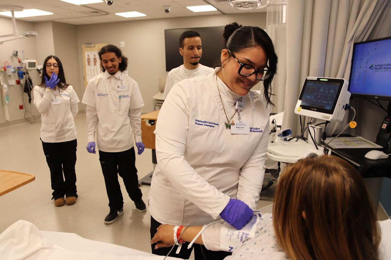 A nursing student in a white coat and purple gloves tends to a patient in a hospital room. Three medical students in uniform observe, smiling, creating a warm and supportive atmosphere.