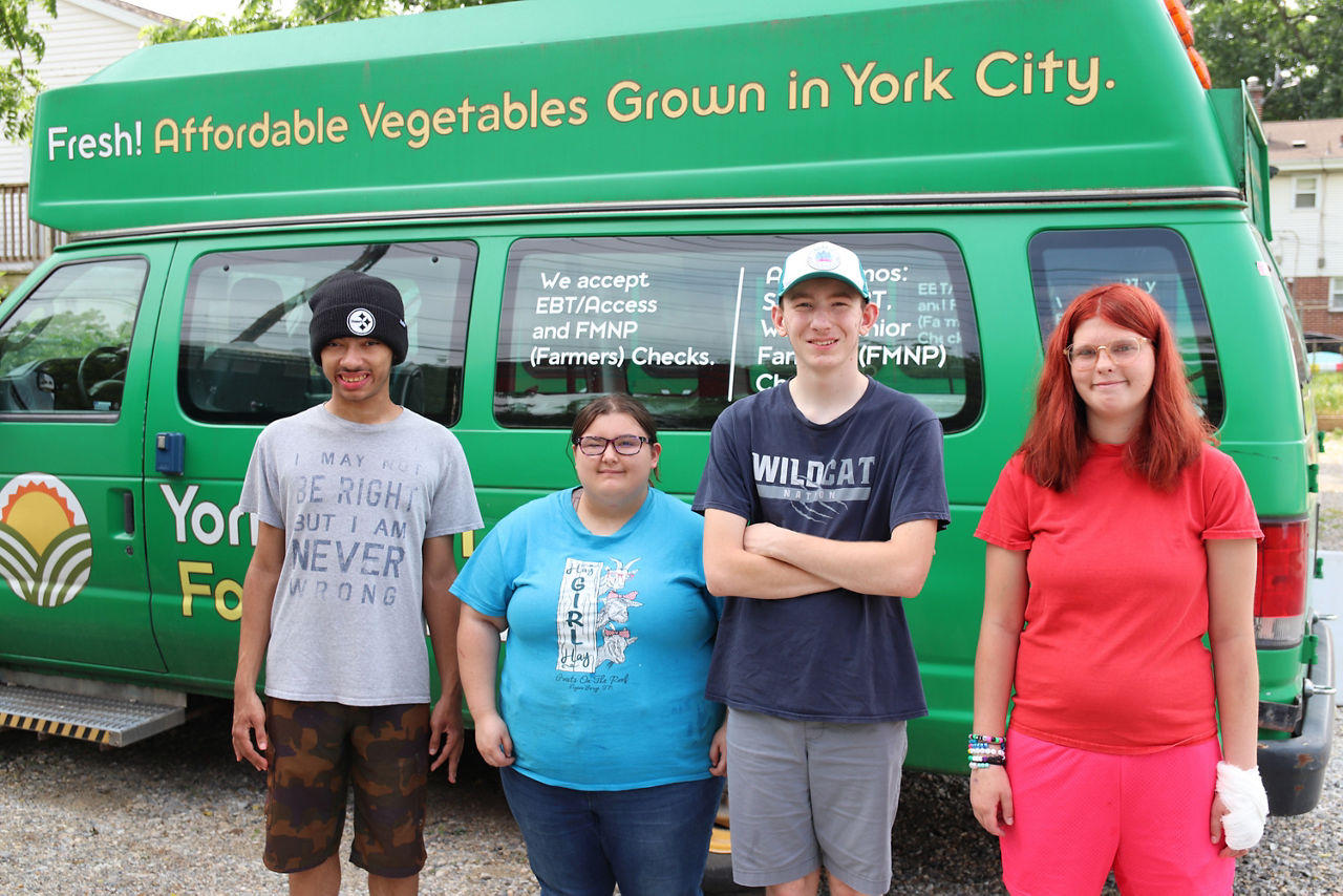 Four people stand smiling in front of a green van that advertises fresh, affordable vegetables grown in York City.