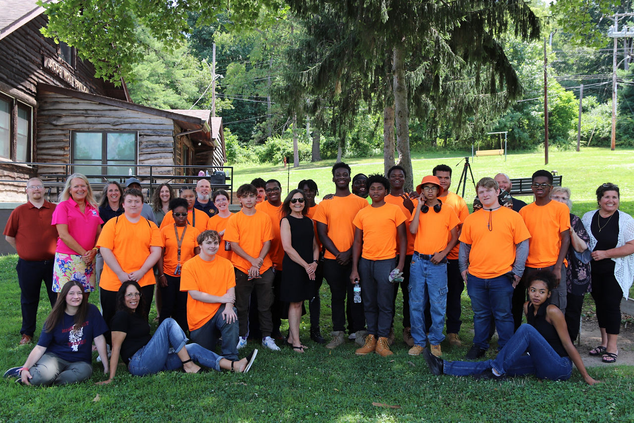 Department of Labor and Industry Secretary Nancy A. Walker stands with a diverse group of staff and students standing on grass, most wearing bright orange shirts, smiling under a tree near a wooden building, conveying teamwork and community.