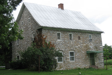 Two story stone house with a center chimney and entrance door on the right.