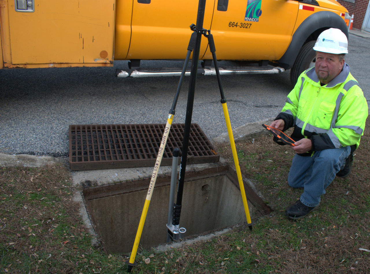 A man wearing a green safety vest and white hard hat holds a computer tablet while kneeling next to an open stormwater drain. An inspection camera sits inside the stormwater drain. A yellow truck sits in the background.