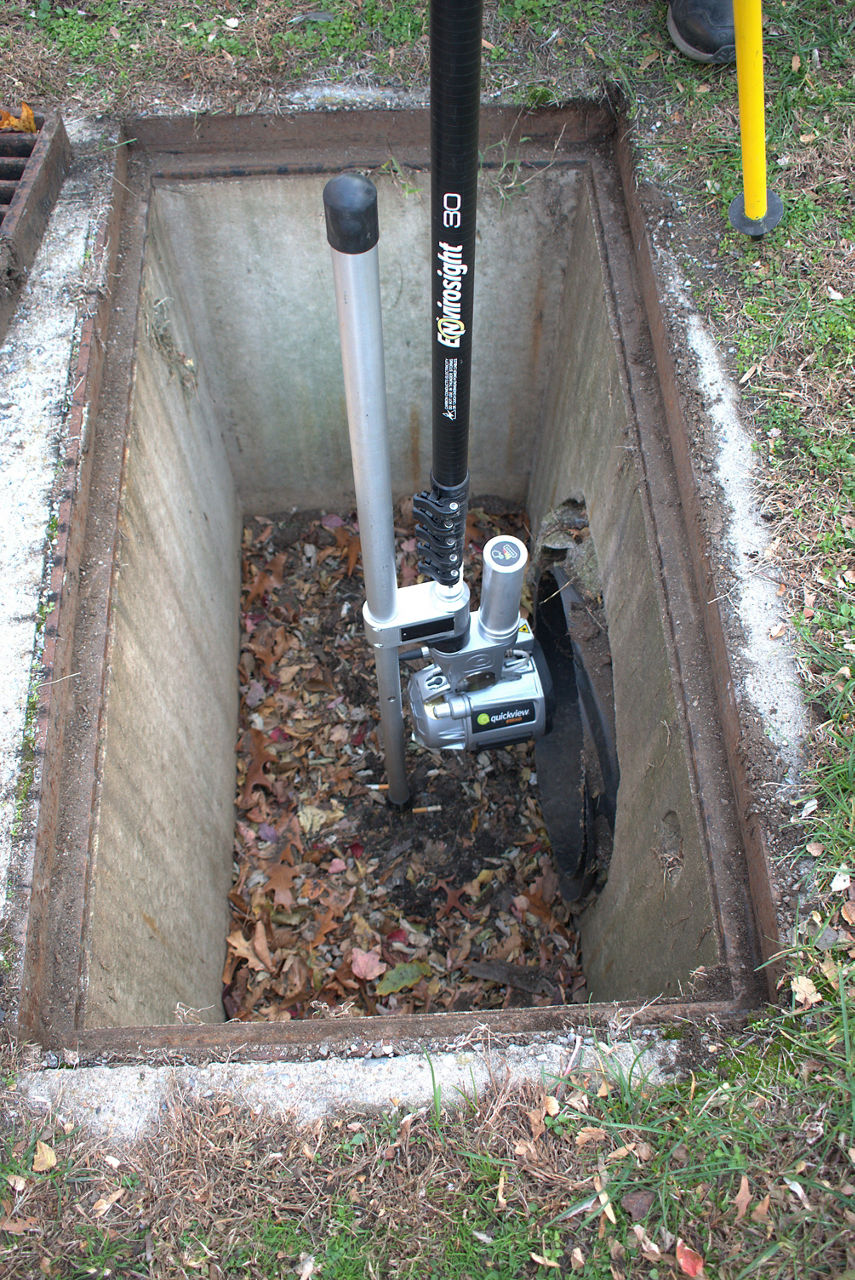 An inspection camera sits inside an open stormwater drain. Green grass is visible around the open stormwater drain.