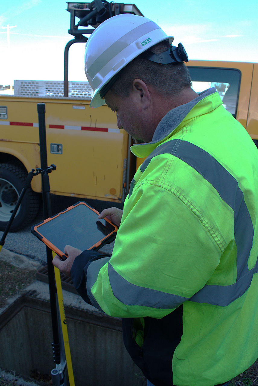 A man wearing a green safety vest and white hard hat holds a computer tablet while kneeling next to an open stormwater drain. An inspection camera sits inside the stormwater drain. A yellow truck sits in the background.